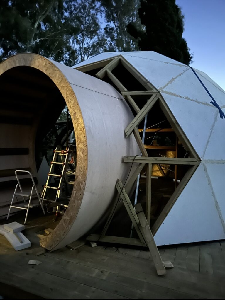 Domo geodésico de madera en fase de construcción con acceso frontal curvo, y esgtructura triangular visible.
