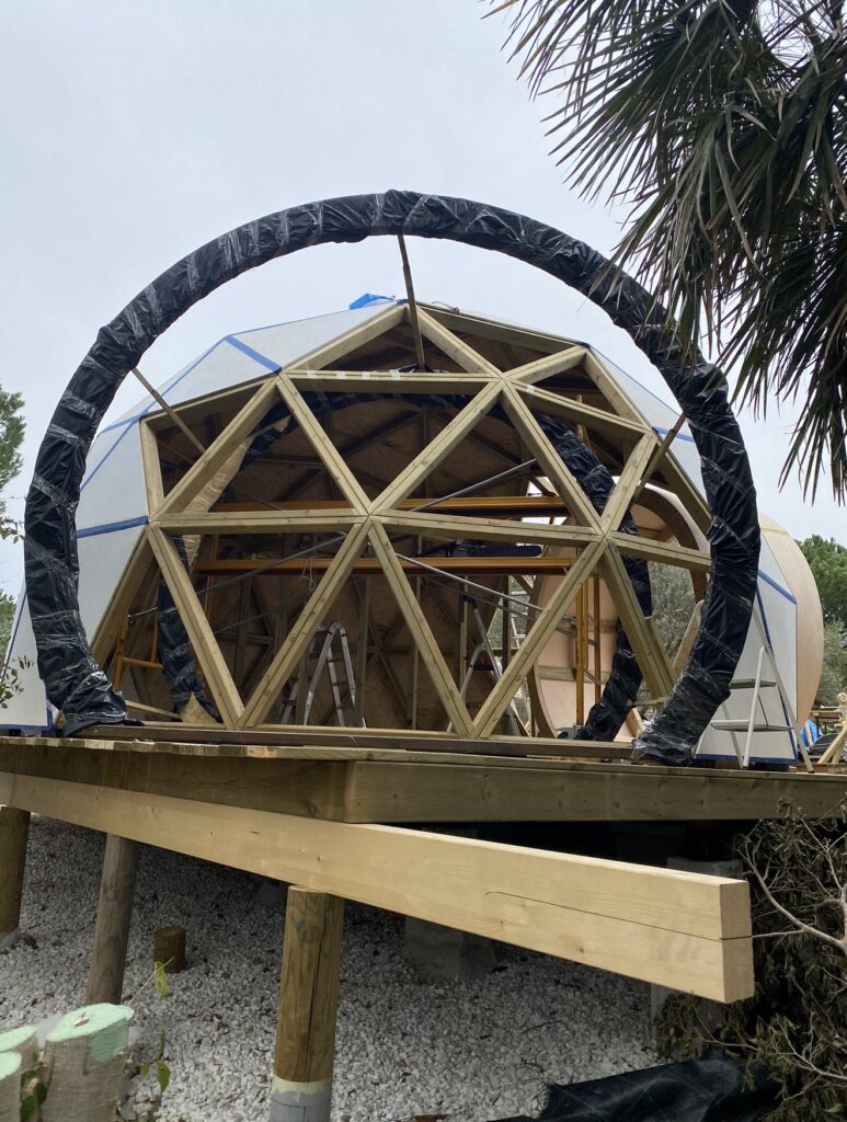 Estructura de domo geodésico de madera en costrucción sobre plataforma elevada, con entramado triangular visible y acceso frontal en arco.